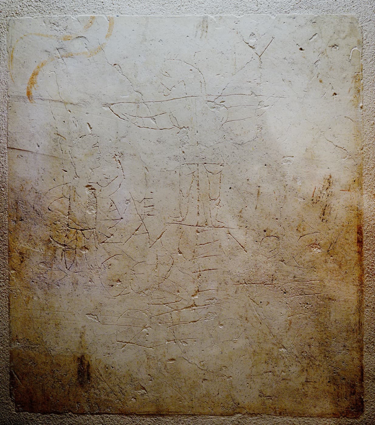 A faint drawing scratched into plaster of a man raising a hand toward a crucified figure with a donkey’s head, with Greek words below