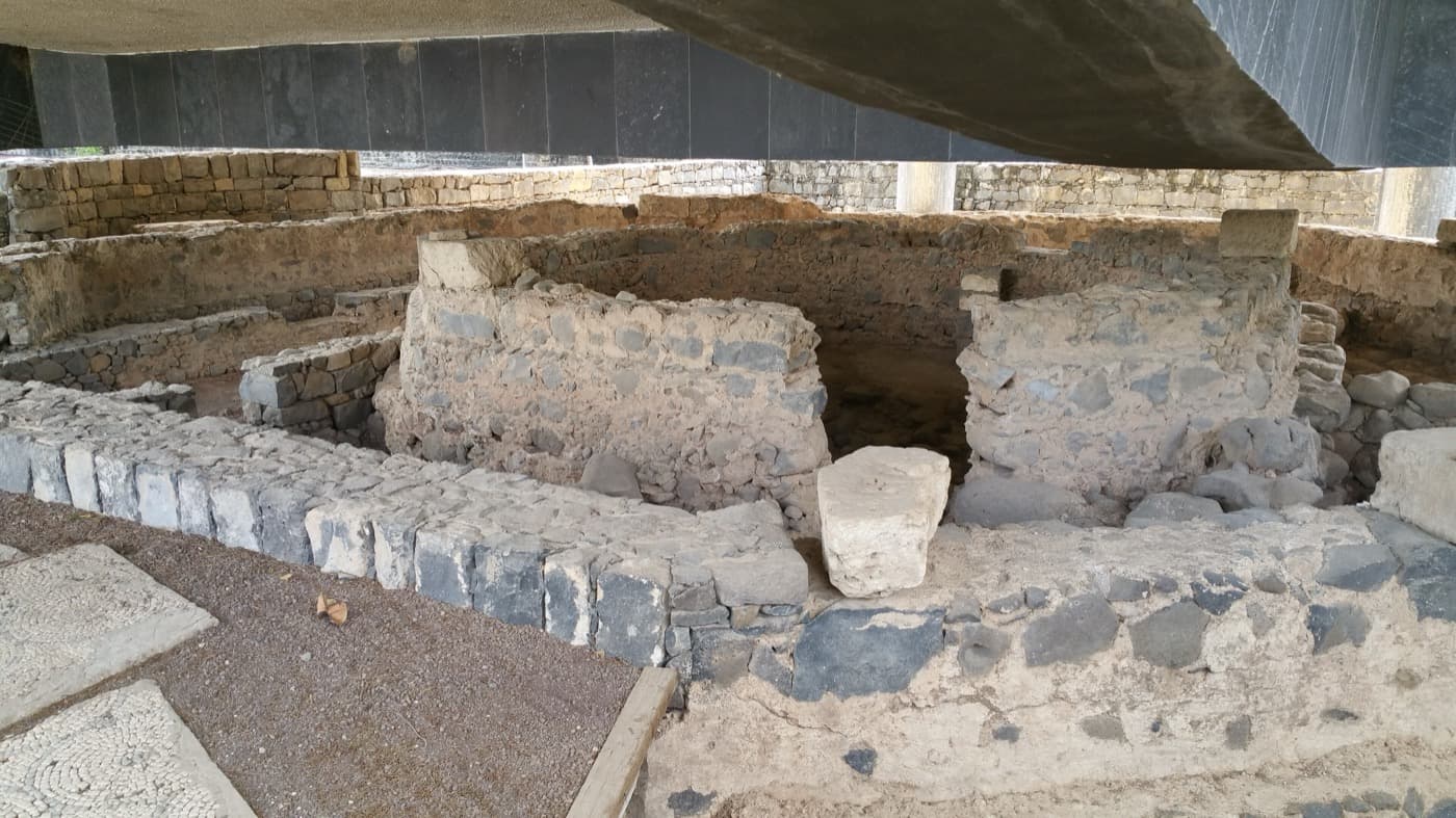 Low black basalt walls of a first-century house inside an octagonal ring of later church walls, seen from above under a modern roof