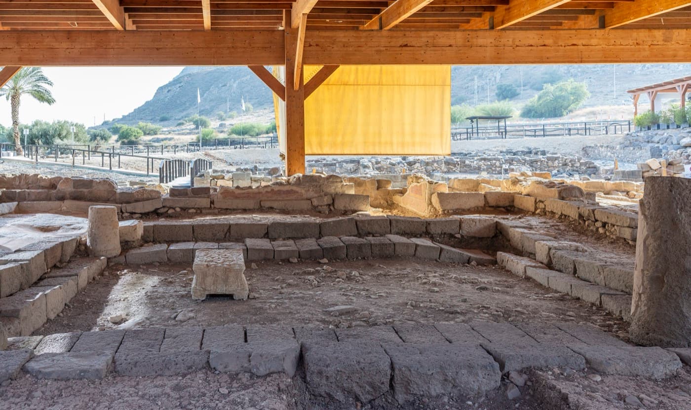 The excavated stone walls and benches of a first-century synagogue under a wooden roof, with a carved stone block in the center